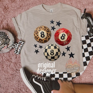 May include: A beige t-shirt featuring three stylized 8-ball designs: leopard print, red with glitter, and black and white checkered. Black star accents surround the balls. The text "original designer" is printed on the shirt.