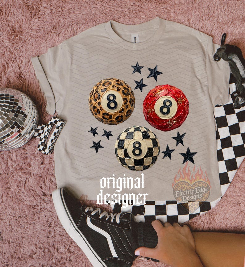 May include: A beige t-shirt featuring three stylized 8-ball designs: leopard print, red with glitter, and black and white checkered. Black star accents surround the balls. The text "original designer" is printed on the shirt.