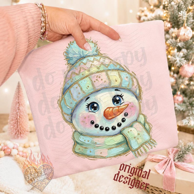May include: A light pink sweatshirt featuring a central illustration of a snowman wearing a pastel-colored knit hat with a pom-pom and a matching scarf. The snowman has a bright orange carrot nose, rosy cheeks, and large blue eyes.