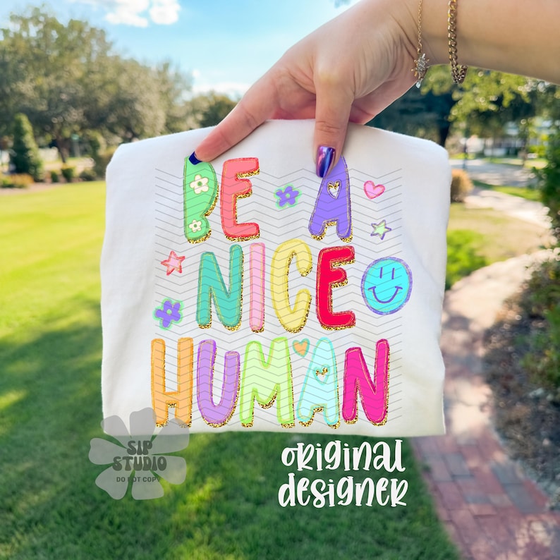 May include: White t-shirt with the phrase "BE A NICE HUMAN" in colorful, glittery letters. The design includes flowers, stars, a heart, and a smiling face. The words "SIP STUDIO" and "original designer" are also printed on the shirt.