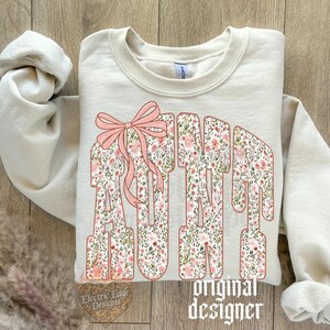 May include: Cream-colored sweatshirt with the word "AUNT" in a floral design, accented with a pink bow. The text "original designer" is printed below the word "AUNT".
