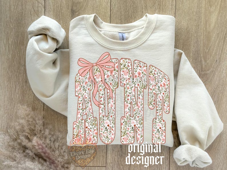 May include: Cream-colored sweatshirt with the word "AUNT" in a floral design, accented with a pink bow. The text "original designer" is printed below the word "AUNT".