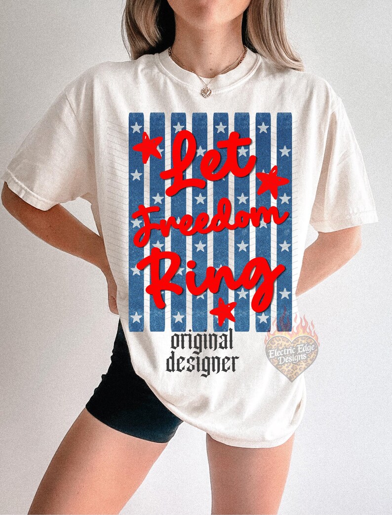 May include: Cream-colored t-shirt with a patriotic design. The graphic features the words "Let Freedom Ring" in red script over a blue and white striped background with stars. The words "original designer" are printed below.