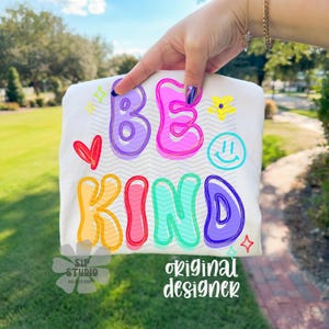 May include: White t-shirt with the words "BE KIND" in colorful bubble letters. The design includes a red heart, a yellow flower, a smiley face, and star accents. The words "SIP STUDIO DO NOT COPY" and "original designer" are also printed on the shirt.