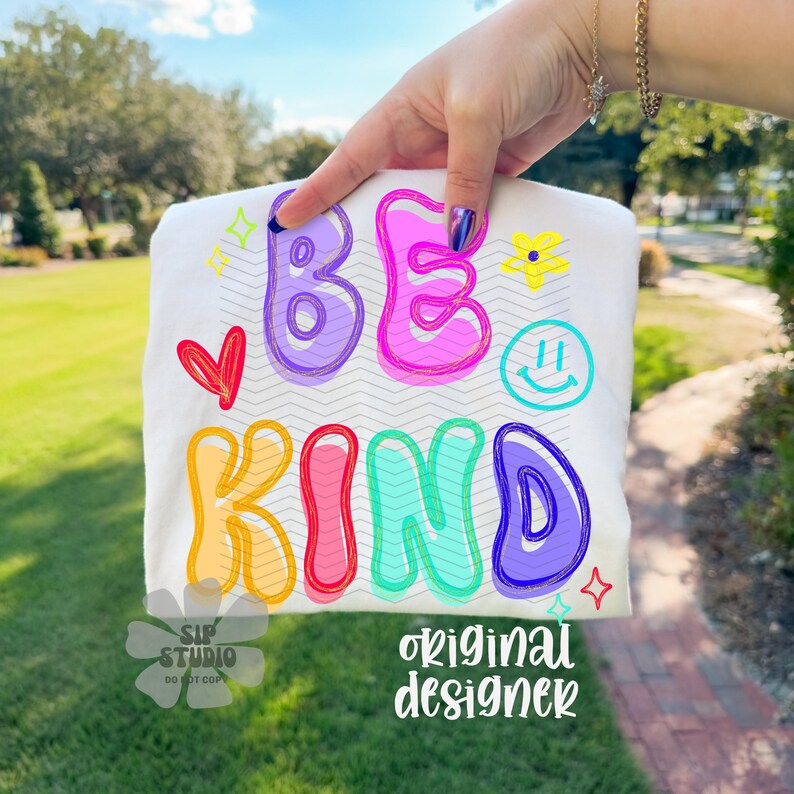 May include: White t-shirt with the words "BE KIND" in colorful bubble letters. The design includes a red heart, a yellow flower, a smiley face, and star accents. The words "SIP STUDIO DO NOT COPY" and "original designer" are also printed on the shirt.