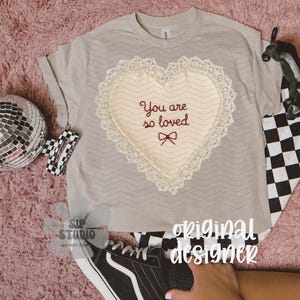 May include: A pale grey t-shirt featuring a heart-shaped design. The heart is cream-coloured with a lace trim and the embroidered text "You are so loved". A black and white chequered pattern is visible.