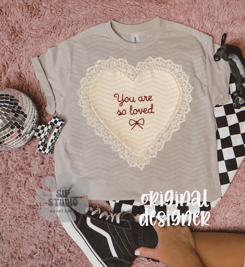 May include: A pale grey t-shirt featuring a heart-shaped design. The heart is cream-coloured with a lace trim and the embroidered text "You are so loved". A black and white chequered pattern is visible.