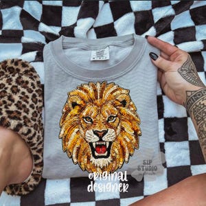 May include: A light gray sweatshirt featuring a detailed, sequined lion's head design in shades of gold, orange, and brown. The text "original designer" is printed at the bottom. The sweatshirt is on a black and white checkered blanket.