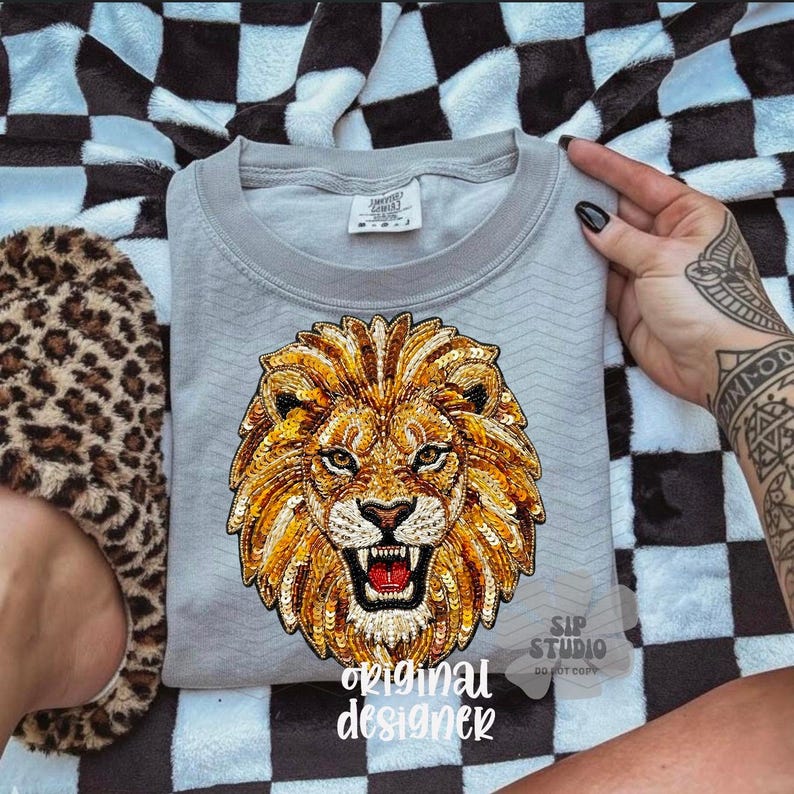 May include: A light gray sweatshirt featuring a detailed, sequined lion's head design in shades of gold, orange, and brown. The text "original designer" is printed at the bottom. The sweatshirt is on a black and white checkered blanket.