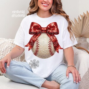 May include: White t-shirt featuring a baseball design. The baseball is adorned with a red sequined bow. The baseball has red stitching. The words "original designer" are printed on the top left of the shirt.