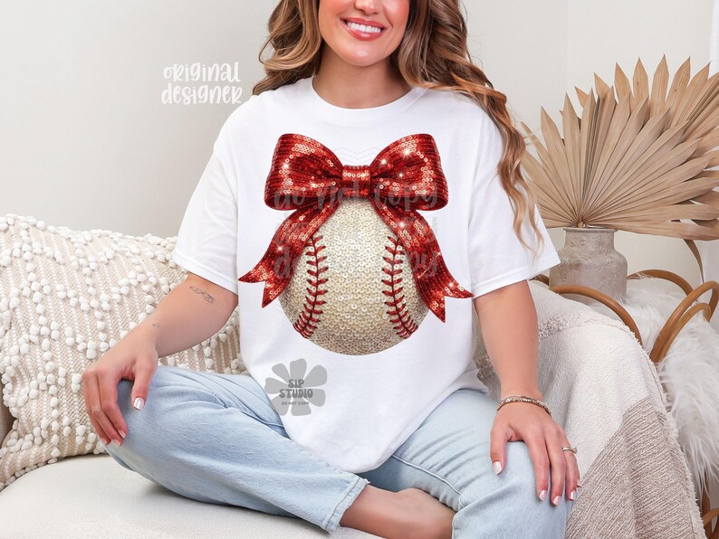 May include: White t-shirt featuring a baseball design. The baseball is adorned with a red sequined bow. The baseball has red stitching. The words "original designer" are printed on the top left of the shirt.