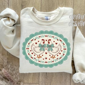 May include: Cream-colored sweatshirt featuring an oval design with two white chickens wearing teal bows. The design is framed by a teal scalloped border with red hearts and bows. The words "original designer" are printed on the top right.