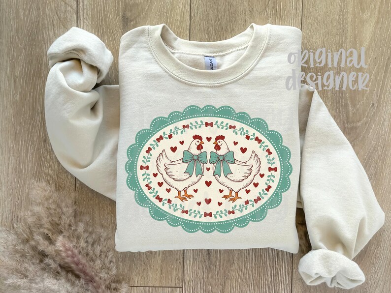 May include: Cream-colored sweatshirt featuring an oval design with two white chickens wearing teal bows. The design is framed by a teal scalloped border with red hearts and bows. The words "original designer" are printed on the top right.