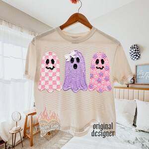 May include: A beige short-sleeve t-shirt featuring a graphic of three ghosts. The left ghost has a pink and white checkerboard pattern, the middle ghost is solid purple with a white bow, and the right ghost is pink with small purple flowers.