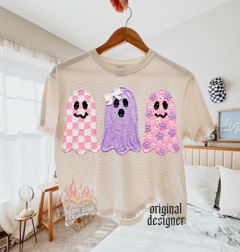 May include: A beige short-sleeve t-shirt featuring a graphic of three ghosts. The left ghost has a pink and white checkerboard pattern, the middle ghost is solid purple with a white bow, and the right ghost is pink with small purple flowers.