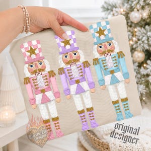 May include: A beige fabric item featuring a printed illustration of three nutcracker figures in pink, lavender, and light blue, each wearing a matching top hat with a gold star, gold epaulets, and gold boots. The text "original designer" is printed in black.