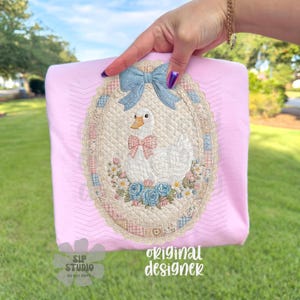 May include: Pink t-shirt featuring an embroidered oval design with a white goose wearing a pink bow tie, surrounded by flowers and a blue bow. The text "original designer" is visible.