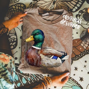 May include: A brown t-shirt featuring a detailed illustration of a mallard duck. The duck has a green head, yellow beak, and colorful plumage. The words "original designer" are printed on the shirt.
