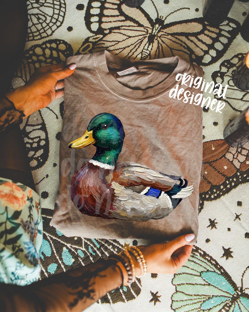 May include: A brown t-shirt featuring a detailed illustration of a mallard duck. The duck has a green head, yellow beak, and colorful plumage. The words "original designer" are printed on the shirt.
