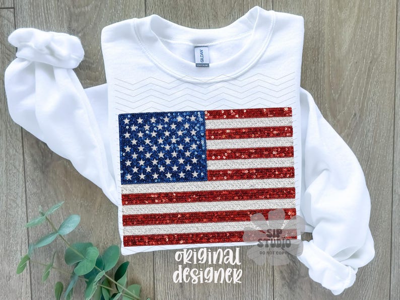 May include: White long-sleeve sweatshirt featuring a sequined American flag design. The flag has a blue square with white stars and red and silver stripes. The words "original designer" are printed at the bottom.