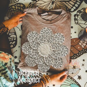 May include: A brown t-shirt with a large, leopard print flower applique. The flower has a ruffled center and is surrounded by petals with a spotted pattern. The words "original designer" are printed on the bottom left of the image.