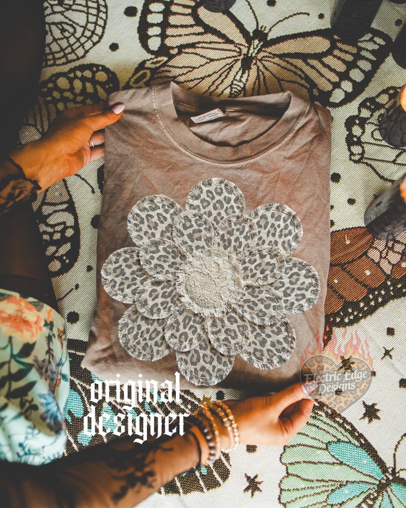 May include: A brown t-shirt with a large, leopard print flower applique. The flower has a ruffled center and is surrounded by petals with a spotted pattern. The words "original designer" are printed on the bottom left of the image.