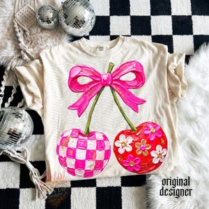 May include: A cream-colored t-shirt featuring a large graphic of two cherries connected by a bright pink bow. One cherry has a pink and white checkered pattern, while the other is red with white and pink flowers. The design includes gold glitter accents.