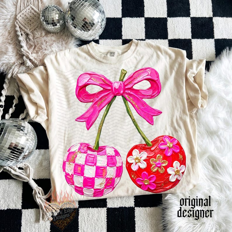 May include: A cream-colored t-shirt featuring a large graphic of two cherries connected by a bright pink bow. One cherry has a pink and white checkered pattern, while the other is red with white and pink flowers. The design includes gold glitter accents.