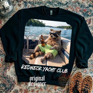 May include: Black sweatshirt featuring a raccoon wearing sunglasses and green striped shorts, holding a beer bottle on a boat. The text "Redneck Yacht Club" and "original designer" are printed on the sweatshirt.