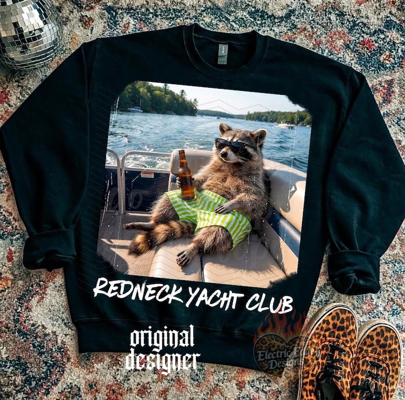 May include: Black sweatshirt featuring a raccoon wearing sunglasses and green striped shorts, holding a beer bottle on a boat. The text "Redneck Yacht Club" and "original designer" are printed on the sweatshirt.