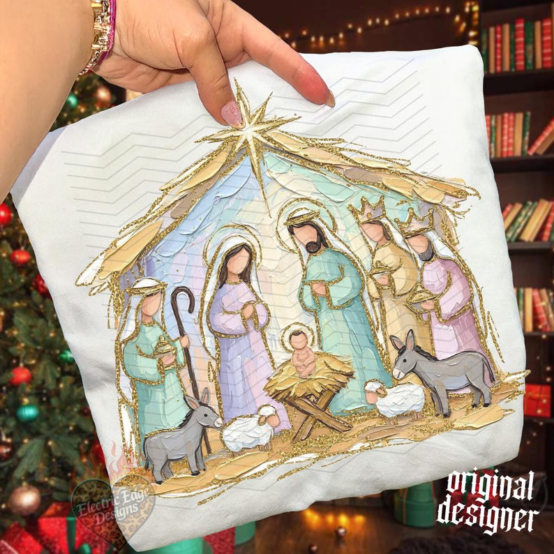 May include: A white fabric item featuring a colorful nativity scene illustration with gold glitter accents. The design depicts figures in pastel robes, a manger with a baby, animals, and a star above, all outlined in sparkling gold glitter.