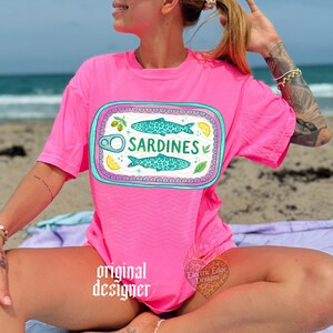 May include: A bright pink t-shirt featuring a sardine can design. The design includes two turquoise fish, the word "SARDINES", and illustrations of lemons and olives. The shirt is a vibrant color and appears to be made of soft material.