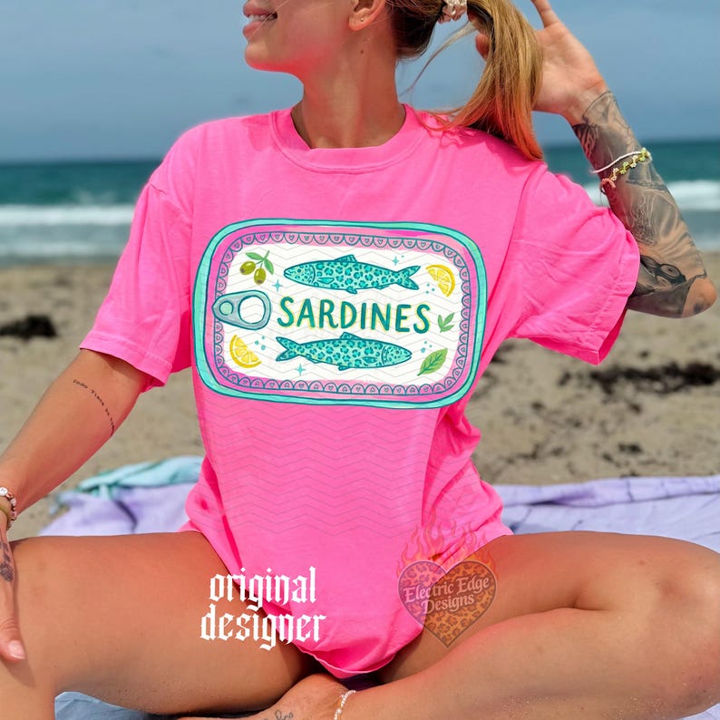 May include: A bright pink t-shirt featuring a sardine can design. The design includes two turquoise fish, the word "SARDINES", and illustrations of lemons and olives. The shirt is a vibrant color and appears to be made of soft material.