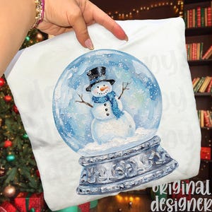 May include: White pillow featuring a snow globe design. The snow globe contains a snowman wearing a black top hat and blue scarf. The base of the snow globe is silver with ornate detailing. The words "original designer" are printed on the bottom right.