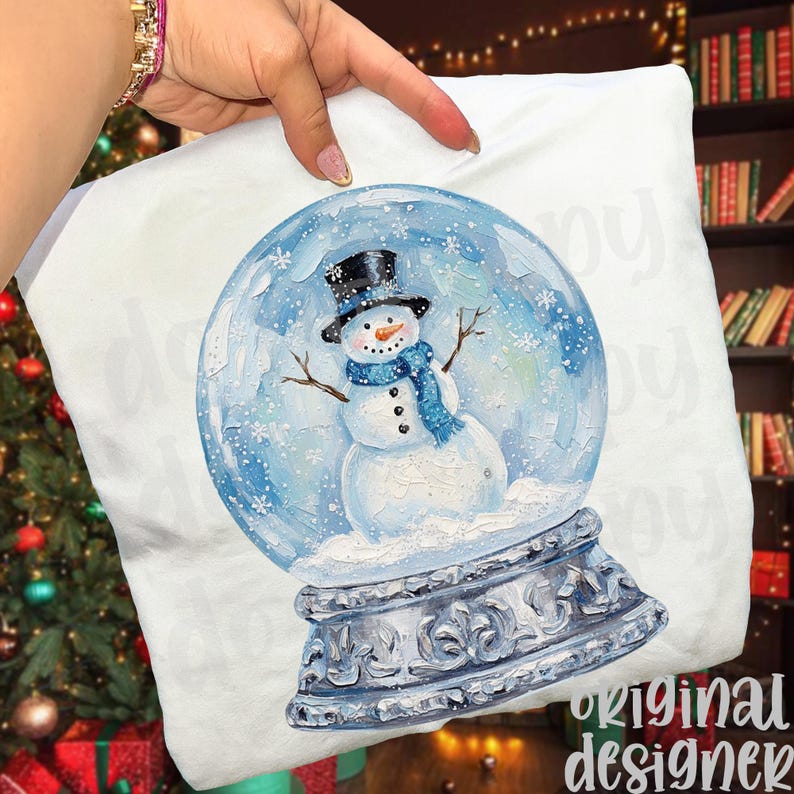 May include: White pillow featuring a snow globe design. The snow globe contains a snowman wearing a black top hat and blue scarf. The base of the snow globe is silver with ornate detailing. The words "original designer" are printed on the bottom right.