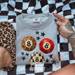 May include: A light gray sweatshirt featuring three 8-ball designs in leopard print, red, and checkered patterns, with black star accents. The text "original designer" is printed at the bottom.