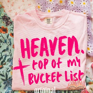 May include: A light pink t-shirt with the words "HEAVEN, top of my BUCKET LIST" in bright pink. The shirt has a zig-zag pattern and a cross graphic. The words "original designer" are printed at the bottom.