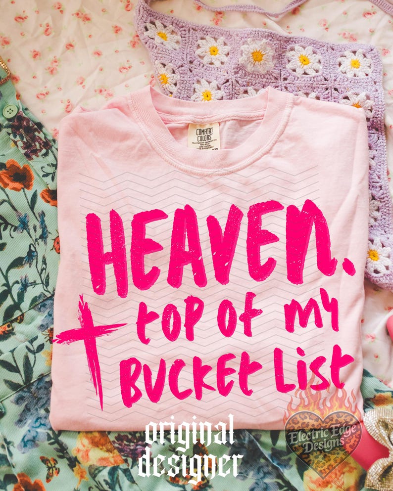 May include: A light pink t-shirt with the words "HEAVEN, top of my BUCKET LIST" in bright pink. The shirt has a zig-zag pattern and a cross graphic. The words "original designer" are printed at the bottom.
