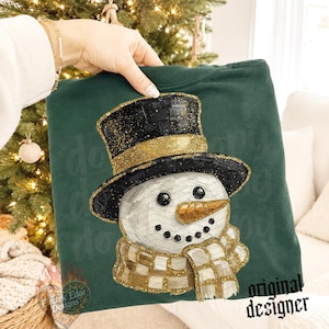 May include: A dark green sweatshirt featuring a graphic of a snowman wearing a black top hat with gold glitter accents. The snowman has a textured white face, a gold glittery carrot nose, and a gold and white checkered scarf with glitter details.