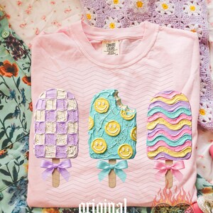 May include: A light pink t-shirt featuring a graphic of three popsicles with textured frosting. The left popsicle has a purple and white checkerboard pattern, the middle is teal with yellow smiley faces, and the right has wavy pastel stripes.
