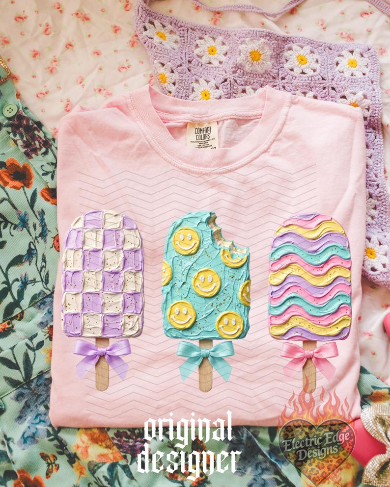 May include: A light pink t-shirt featuring a graphic of three popsicles with textured frosting. The left popsicle has a purple and white checkerboard pattern, the middle is teal with yellow smiley faces, and the right has wavy pastel stripes.