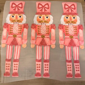 May include: Three identical nutcracker figures printed on a translucent sheet. Each nutcracker features a pink uniform with gold glitter accents, a tall hat with a pink bow, white hair, a gold mustache, and pink striped trousers with glittery boots.