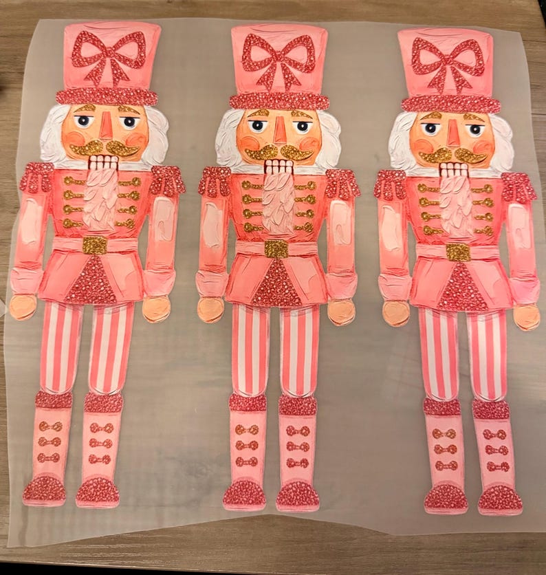 May include: Three identical nutcracker figures printed on a translucent sheet. Each nutcracker features a pink uniform with gold glitter accents, a tall hat with a pink bow, white hair, a gold mustache, and pink striped trousers with glittery boots.