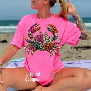 May include: Bright pink t-shirt featuring a crab design with a patchwork pattern in various colors and leopard print. The crab has rhinestone accents. The words "original designer" are printed on the shirt. The shirt is a casual beachwear item.