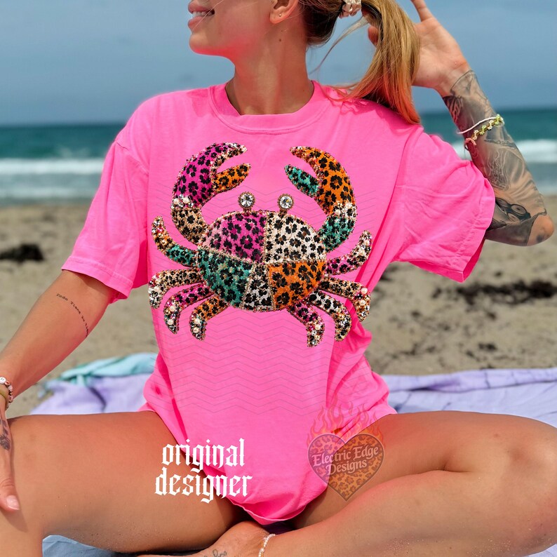 May include: Bright pink t-shirt featuring a crab design with a patchwork pattern in various colors and leopard print. The crab has rhinestone accents. The words "original designer" are printed on the shirt. The shirt is a casual beachwear item.