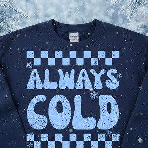 May include: Navy blue sweatshirt with the words "ALWAYS COLD" in light blue, with a checkered pattern above and below the text. The sweatshirt has a crew neck and a tag that says "GILDAN".