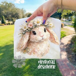 May include: A white t-shirt featuring a watercolor illustration of a bunny wearing a floral crown. The bunny has long ears and big eyes. The text "original designer" is printed on the shirt.