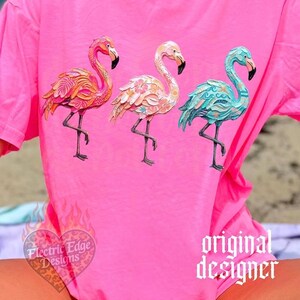 May include: A bright pink t-shirt featuring three flamingo designs in shades of orange, pink, and teal. The flamingos have intricate patterns and stand on one leg. The shirt also has the text "Electric Edge Designs" and "original designer".