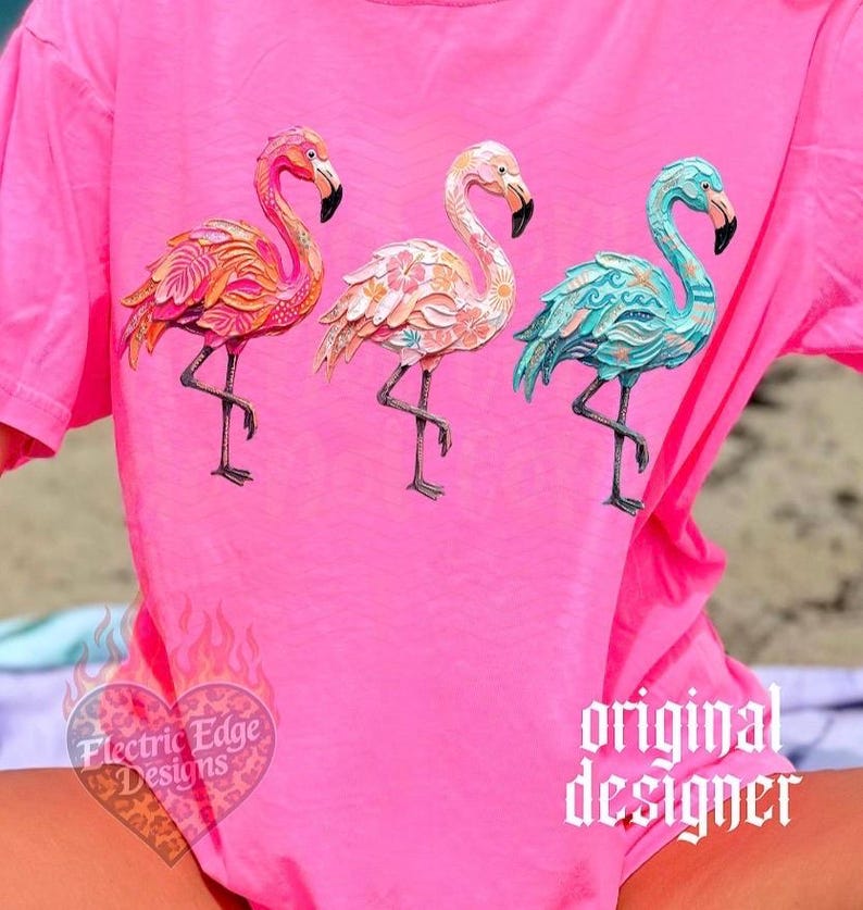May include: A bright pink t-shirt featuring three flamingo designs in shades of orange, pink, and teal. The flamingos have intricate patterns and stand on one leg. The shirt also has the text "Electric Edge Designs" and "original designer".
