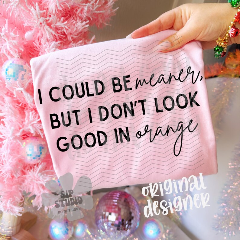 May include: A light pink t-shirt with the text "I COULD BE meaner, BUT I DON'T LOOK GOOD IN orange" in black script. The shirt has a zig-zag pattern and is held up by a hand. The background includes a pink Christmas tree and disco balls.